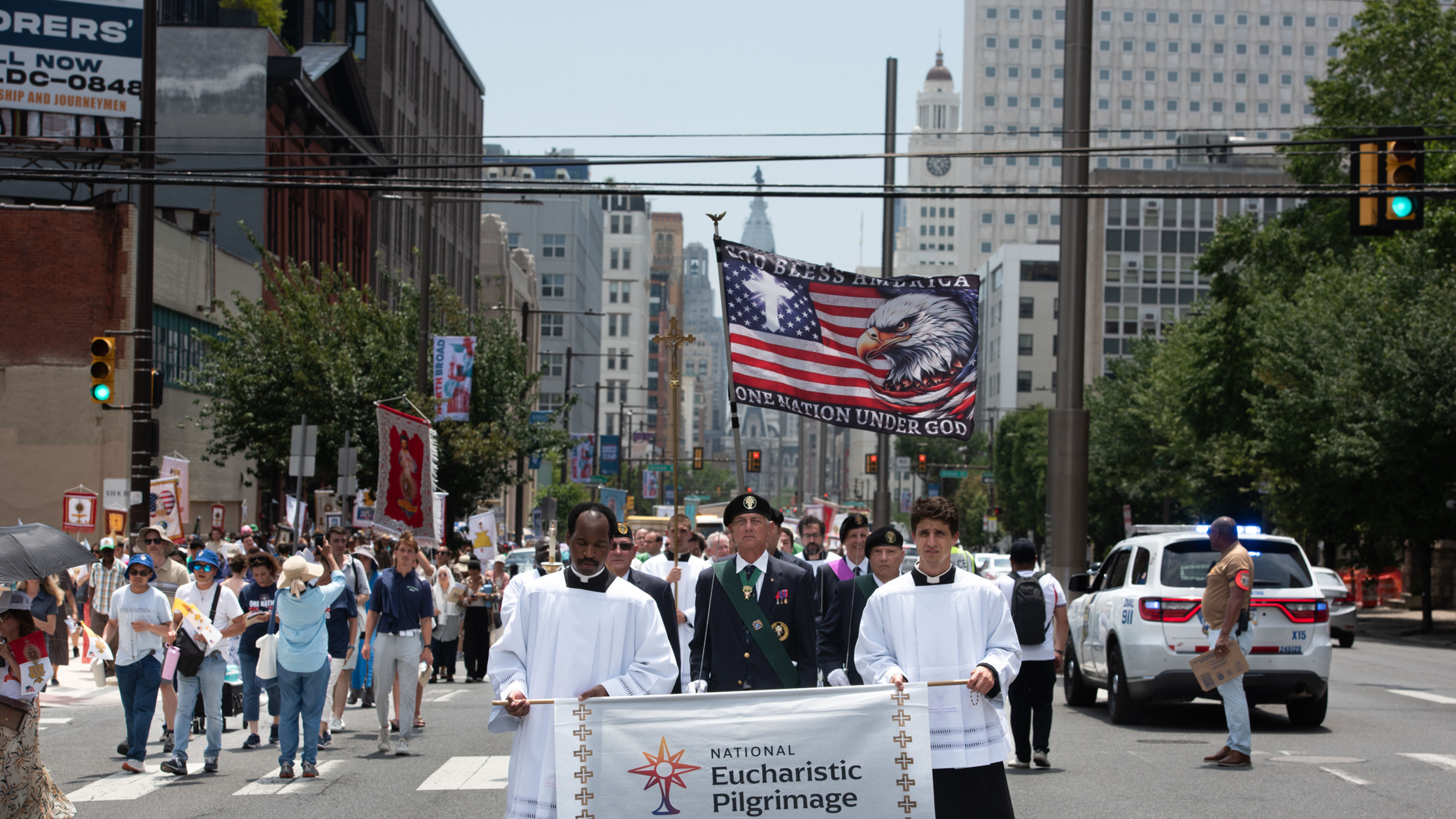 Participants carry the National Eucharistic Pilgrimage banner beneath a flag reading “God Bless America” and “One Nation Under God” during the procession through Philadelphia, Sunday, July 5, 2026. | Credit: Jeffrey Bruno
