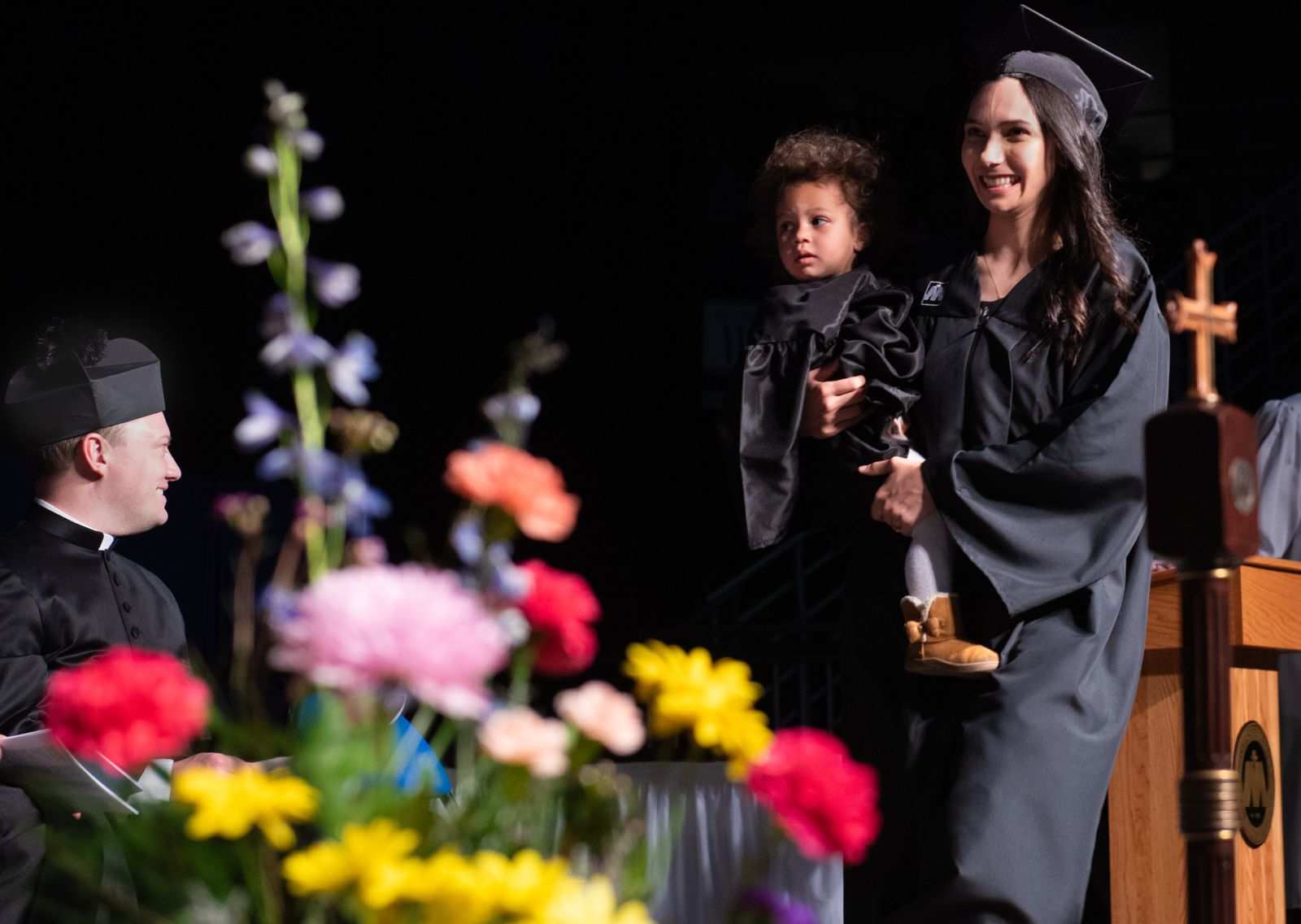 Student mom Katie O'Meara (Chihoski) walks the stage with her toddler, Lucia, by her side at commencement in 2024. Father Dominic Bouck, chaplain at the university, is pictured at left. | Credit: Mike McCleary/University of Mary
