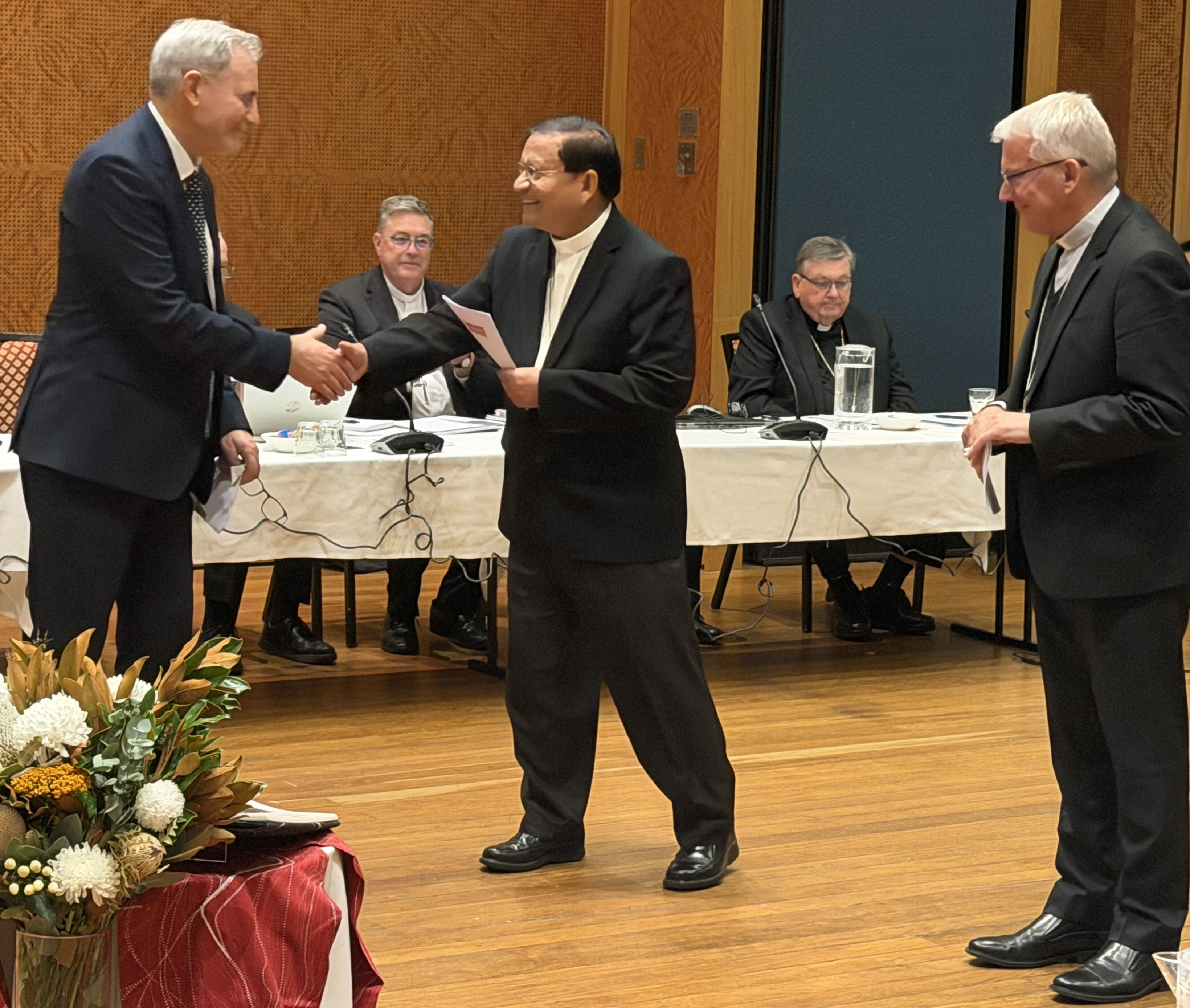 Cardinal Charles Maung Bo (center) hands the commissioning document to Peter Gates (left), the new national director of Catholic Mission Australia, in the presence of Archbishop Timothy Costelloe, SDB (right), president of the Australian Catholic Bishops' Conference, in Sydney on May 8, 2026. | Credit: Paul Osborne/ACBC