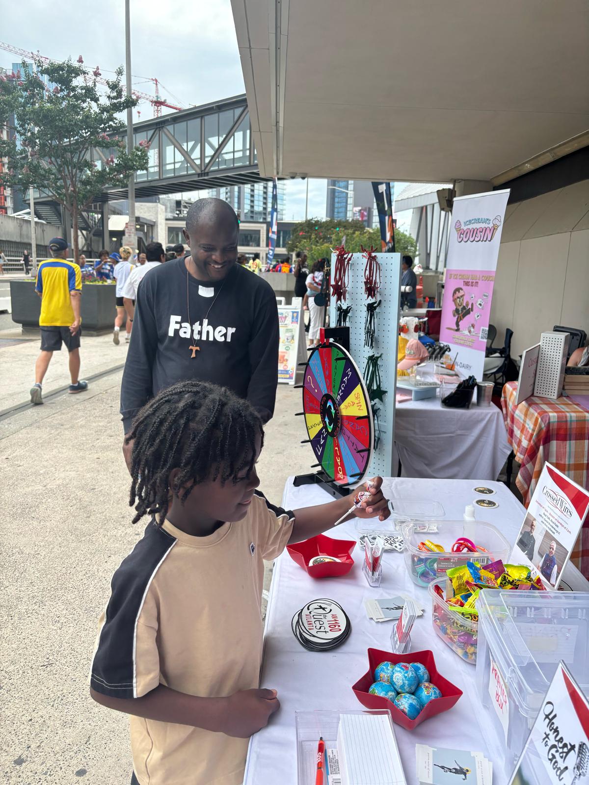 Father Valery Akoh, pastor of St. Matthew Catholic Church in Tyrone, Georgia, talks with a boy at The Quest Atlanta Catholic Radio’s hospitality booth in Atlanta during the World Cup. | Credit: Photo courtesy of The Quest Atlanta Catholic Radio