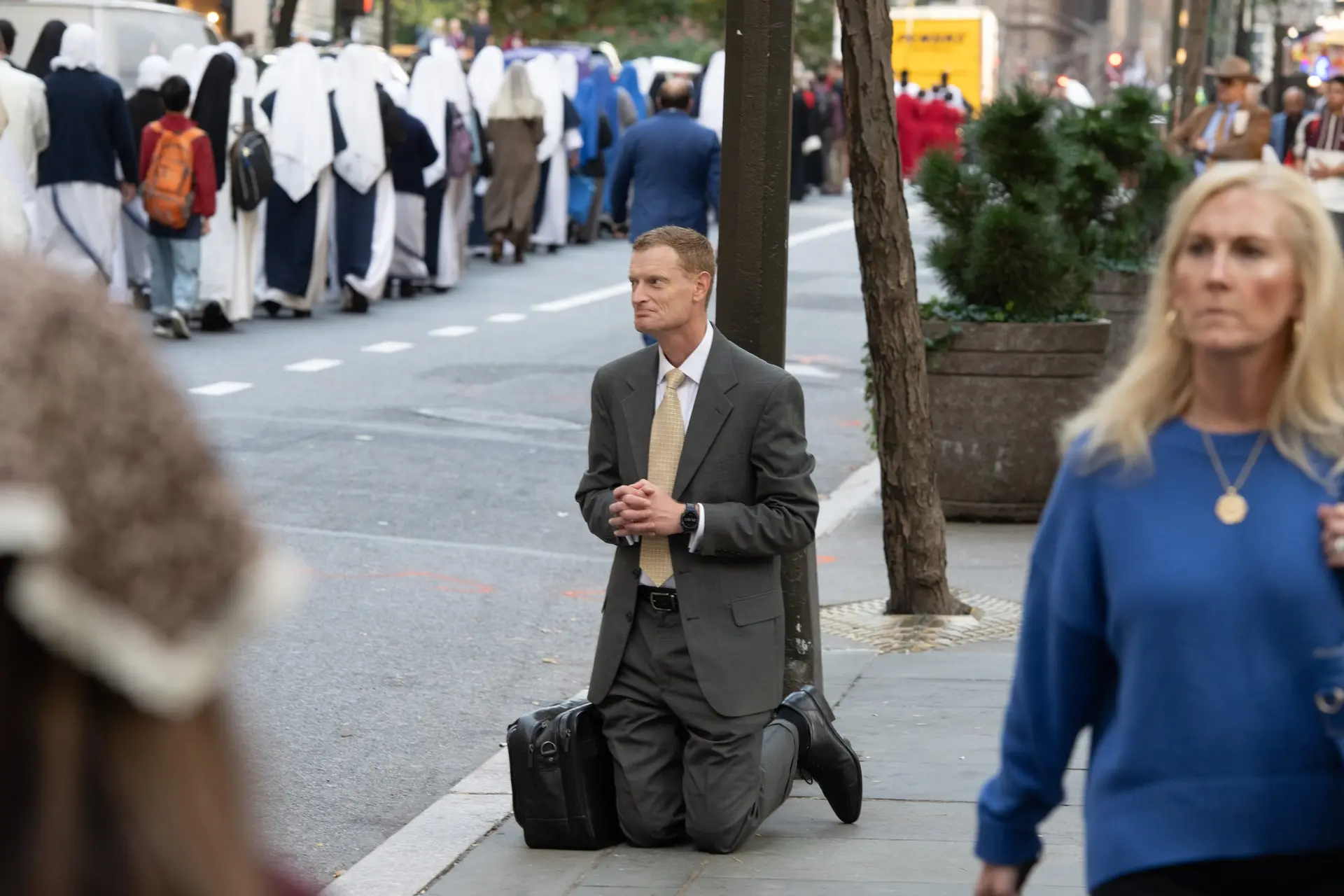 A man kneels on the sidewalk beside his bag as the procession passes. An ordinary New York corner becomes a place of adoration. | Credit: Jeffrey Bruno