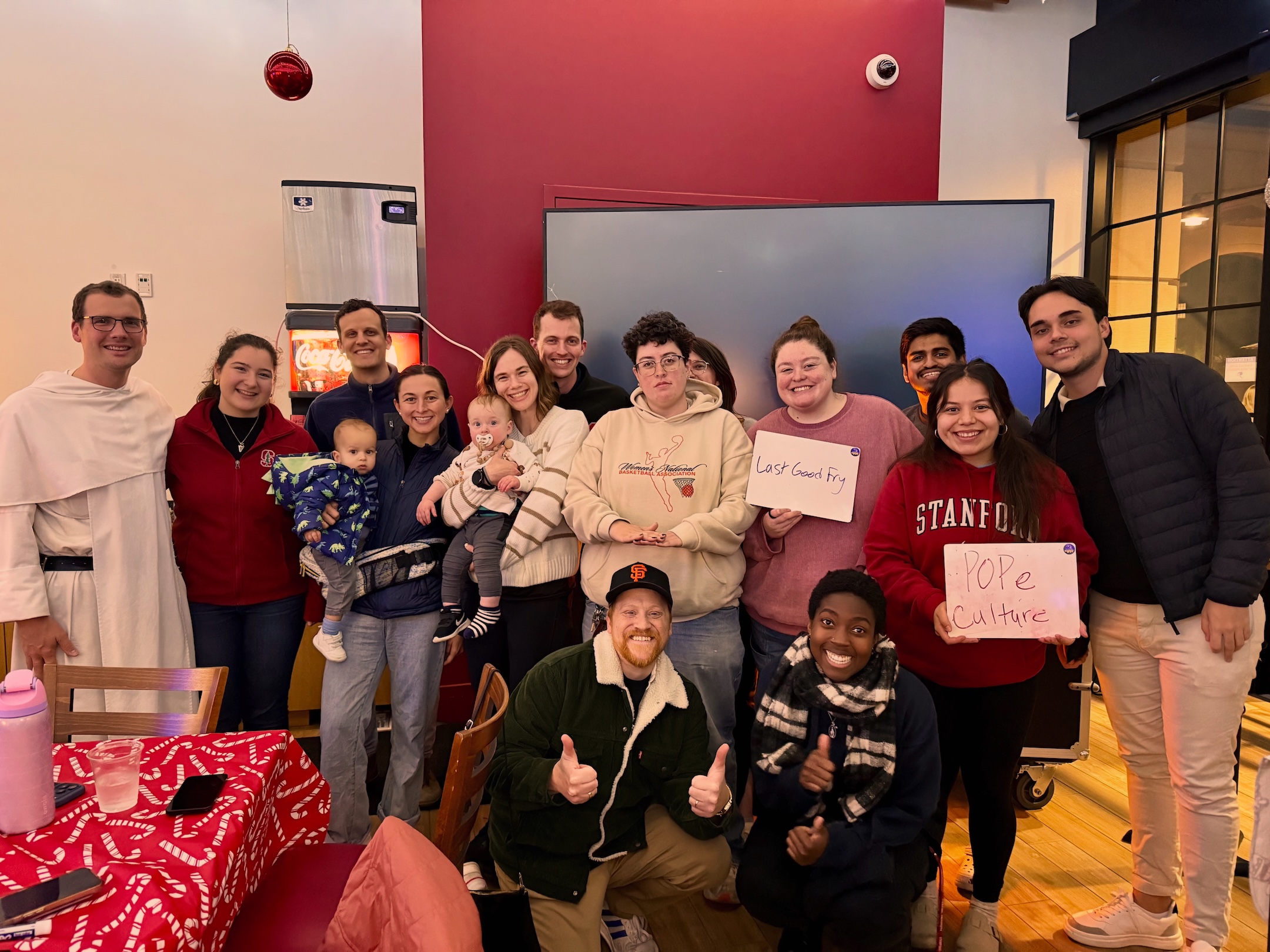 Members of Stanford University's Catholic communuity gather for grad trivia night. From left to right: Father Chrysostom Mijinke, OP,  Isabella McCormick, Andrew Cheshire, Marlena Cheshire holding Michael Cheshire (baby), Caneel Cooper holding Warren Cooper, Matteus Cooper, Unknown, Maria Moore, Nevin Aresh, Lisa Archuleta, Gabriel. Bottom: Bo Lotti, Uwade Akhere. | Photo courtesy of Lisa Archuleta,