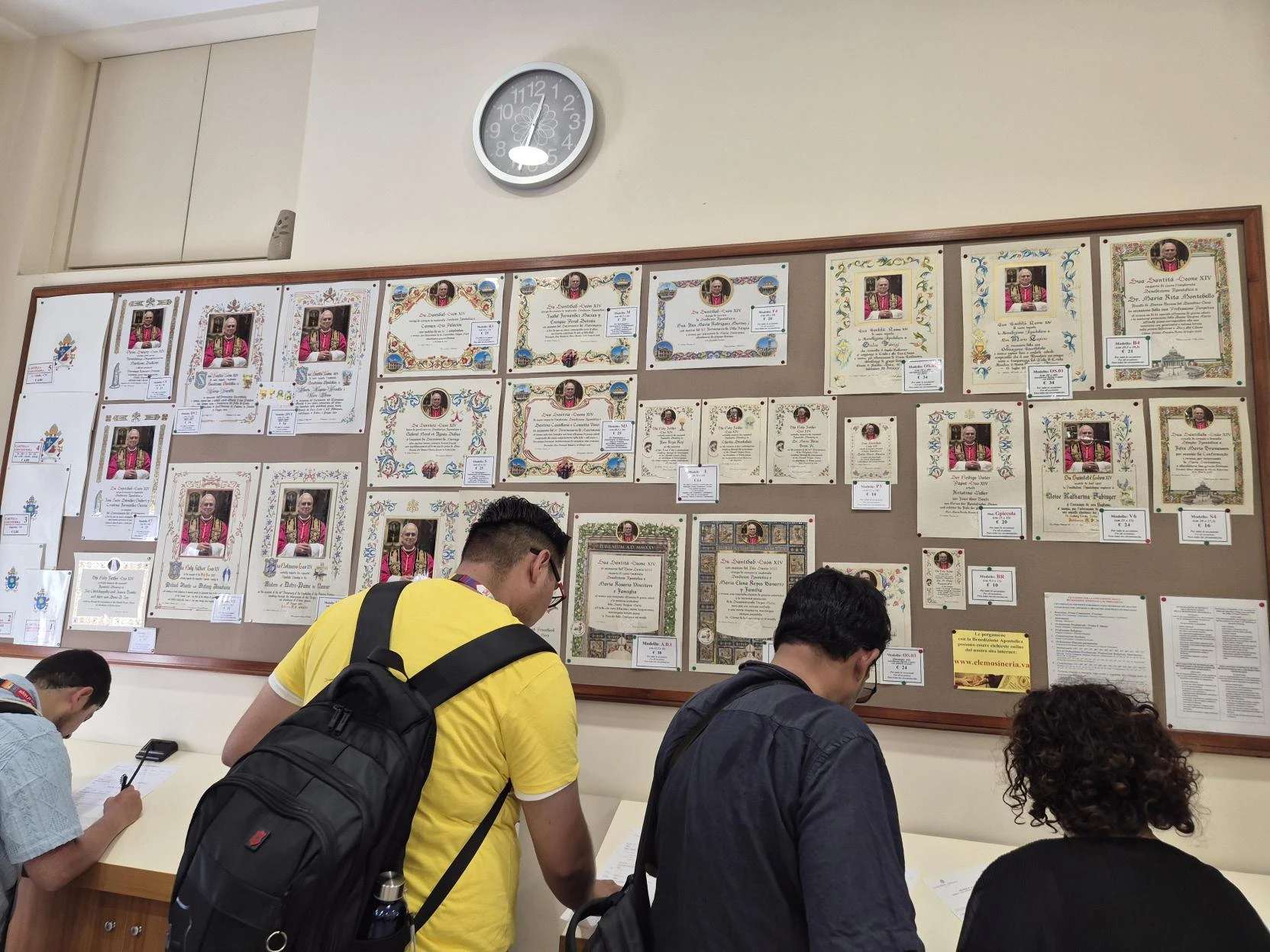 Bezoekers bekijken borden die in het kantoor van de pauselijke liefdadigheid in Vaticaanstad hangen, waarop de verschillende modellen pauselijke zegencertificaten staan die besteld kunnen worden, dinsdag 29 juli 2025. Credit: Hannah Brockhaus/CNA