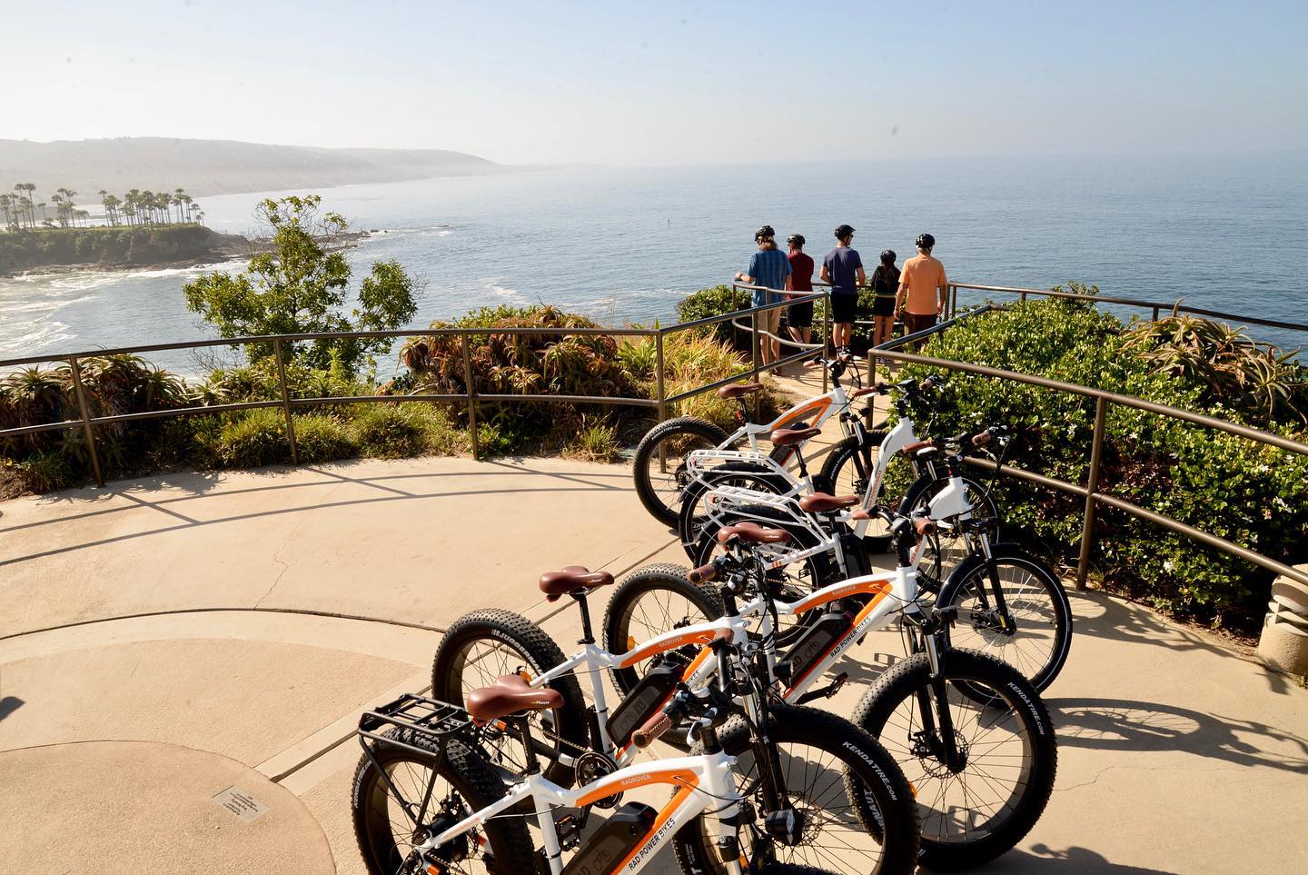 Group of cyclists standing at a scenic overlook in Laguna Beach, with electric bikes parked nearby and a view of the ocean and coastline in the background.