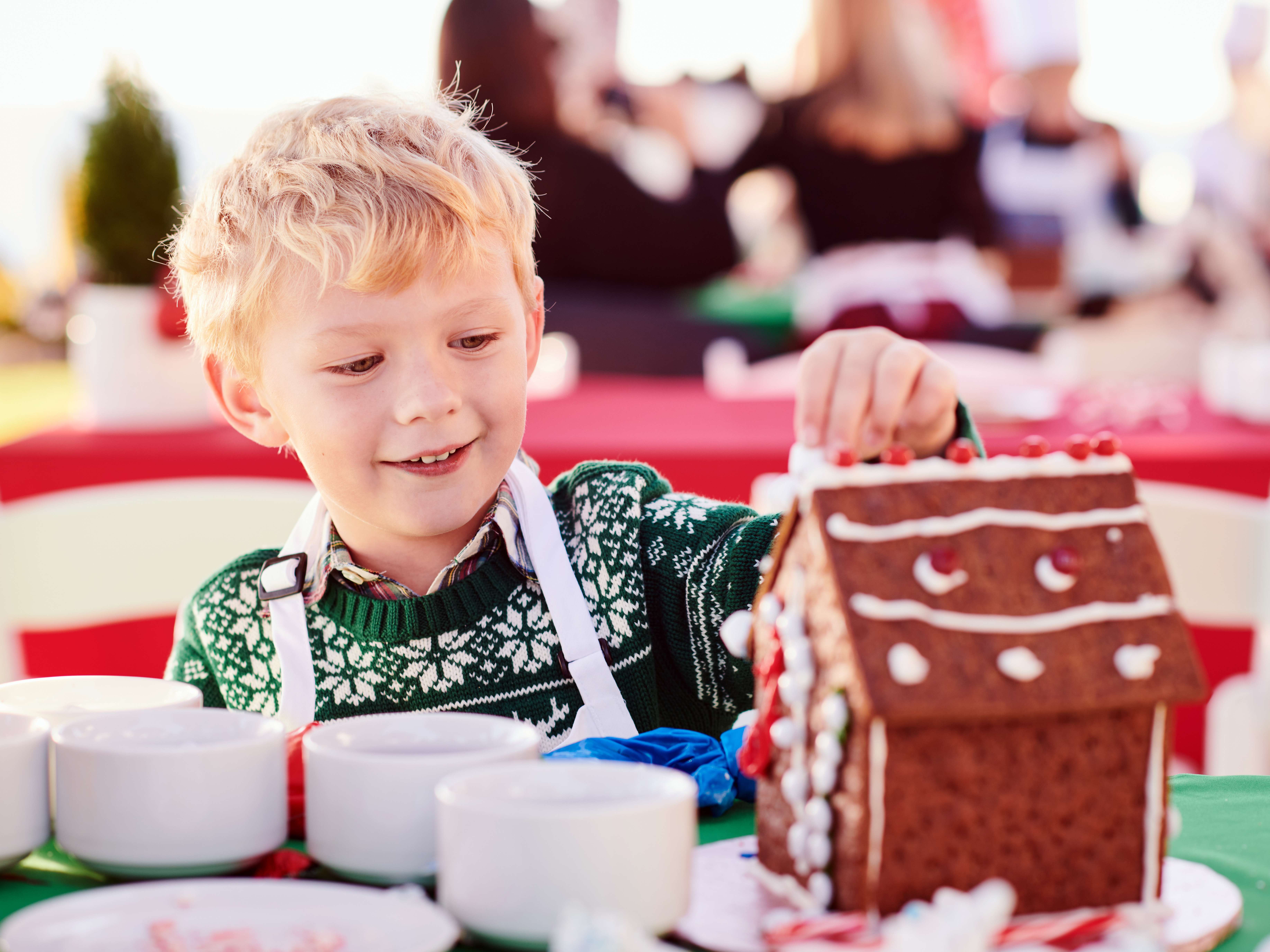 Young boy decorating a gingerbread house during a holiday event at Montage Laguna Beach, with a festive atmosphere in the background.