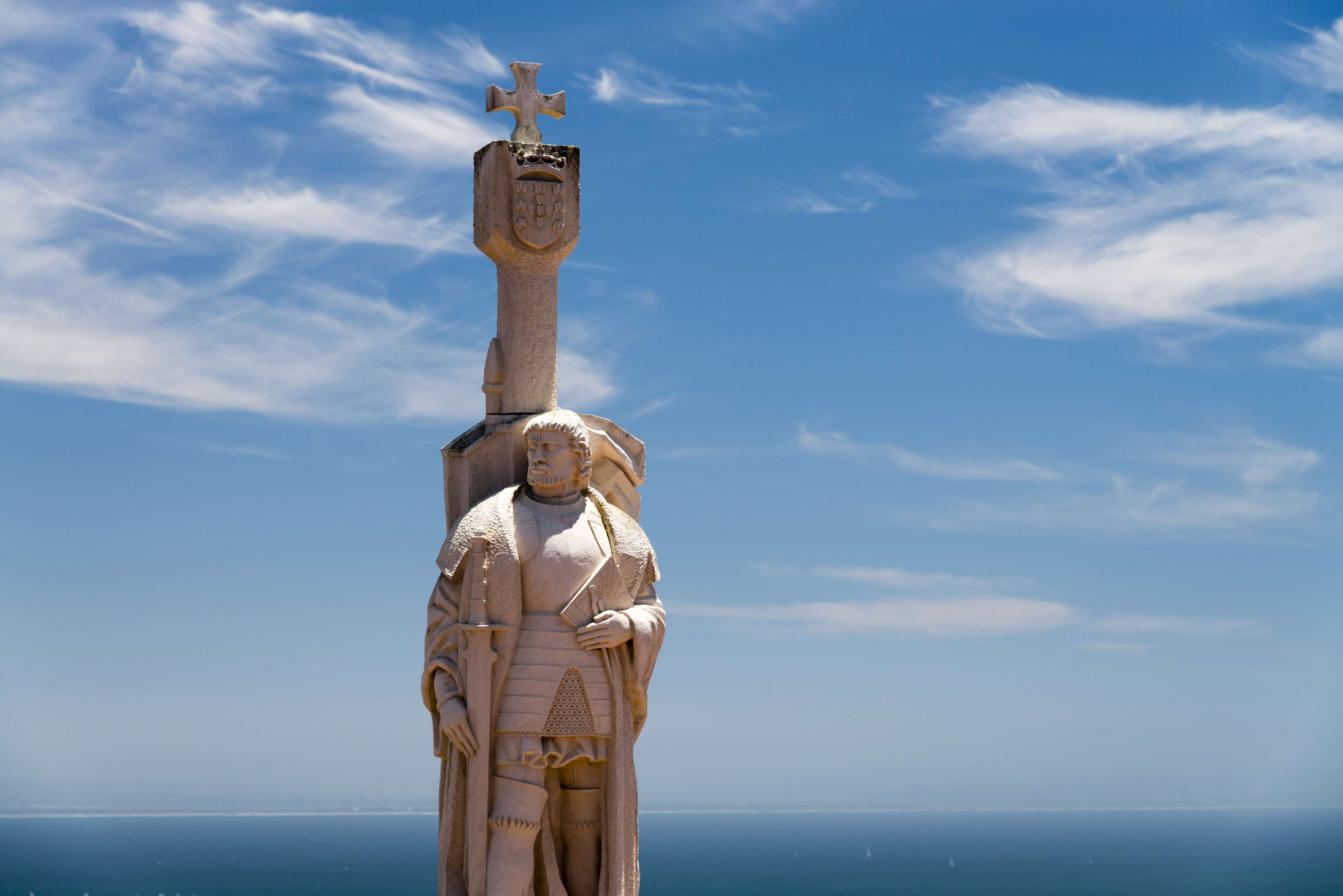 A stone statue of a historical figure in armor, holding a cross, set against a clear blue sky and ocean horizon, with the head obscured by a square block.