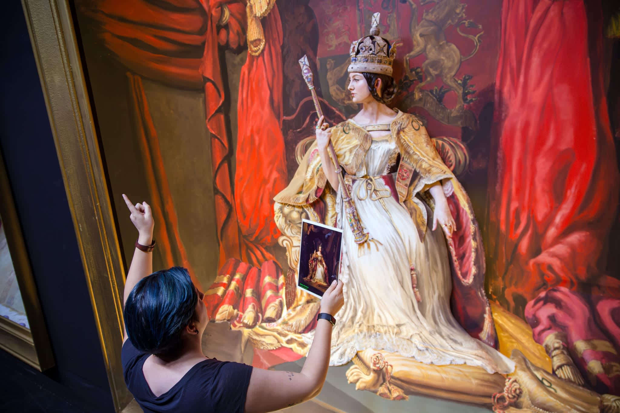 Person observing and pointing at a large, detailed painting of a queen in royal attire, holding a smaller reference image of the same artwork in hand.