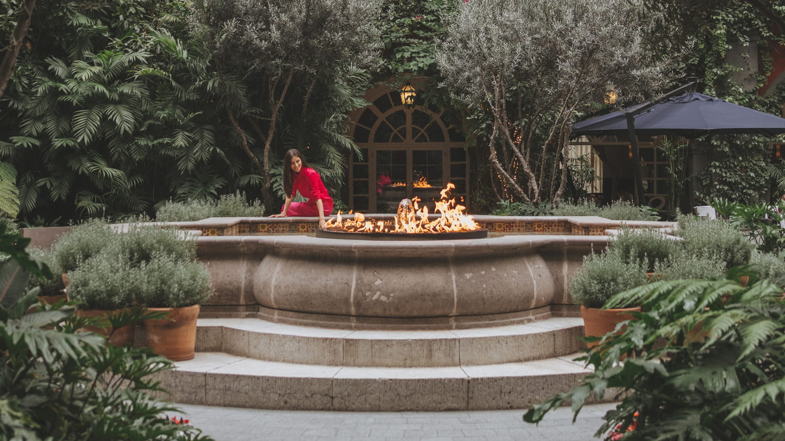 A woman in a red dress sits by a fire pit surrounded by lush greenery in a courtyard.