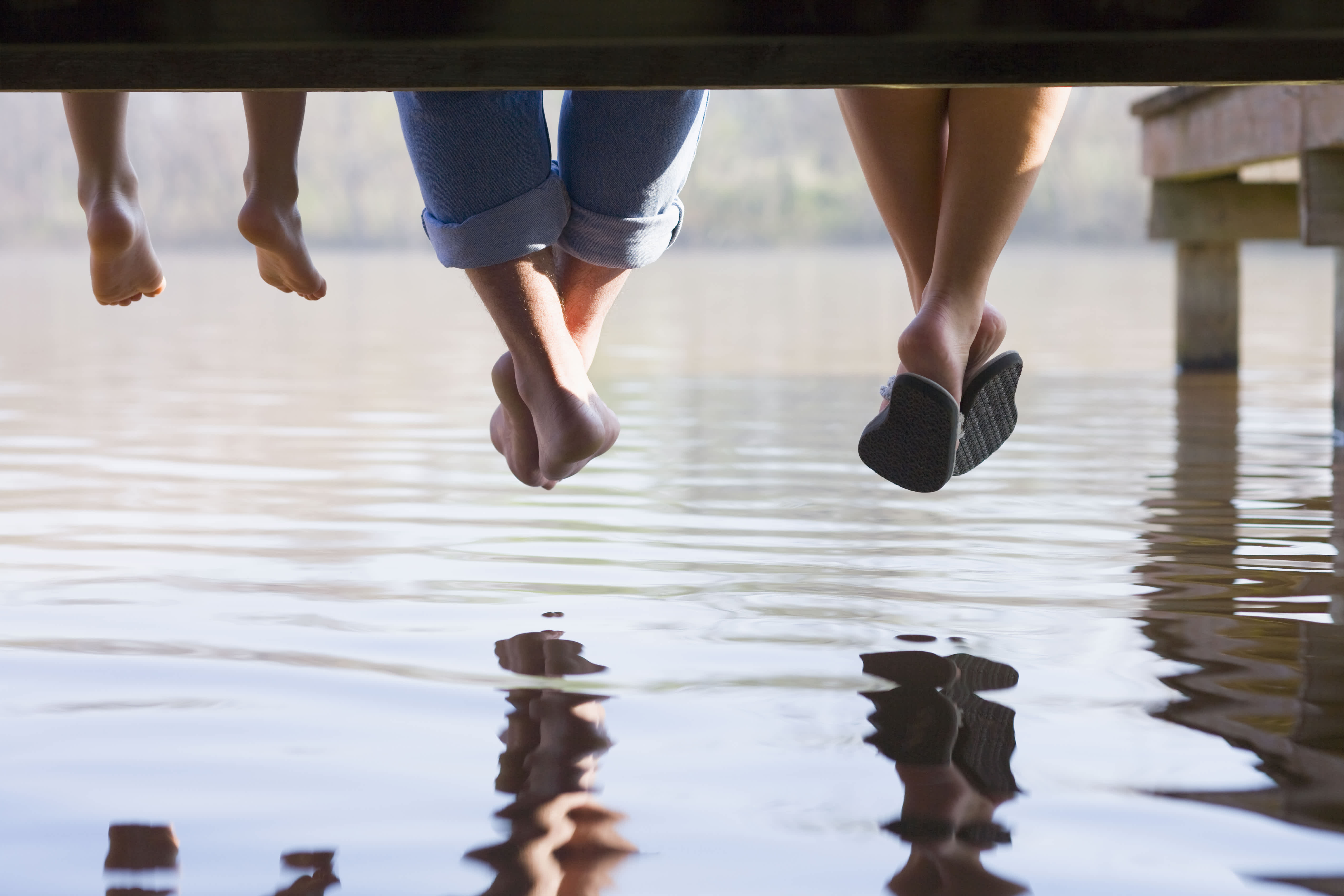 Three people sitting on a dock with their legs dangling over the water, their reflections visible on the calm surface below.