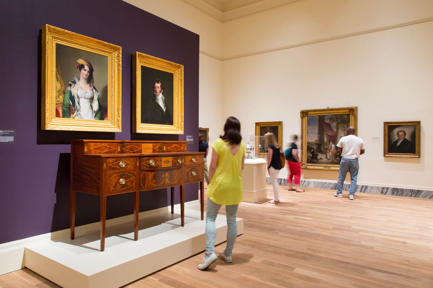 Visitors observing classic portraits and antique furniture in an art gallery at the Gibbes Museum of Art.