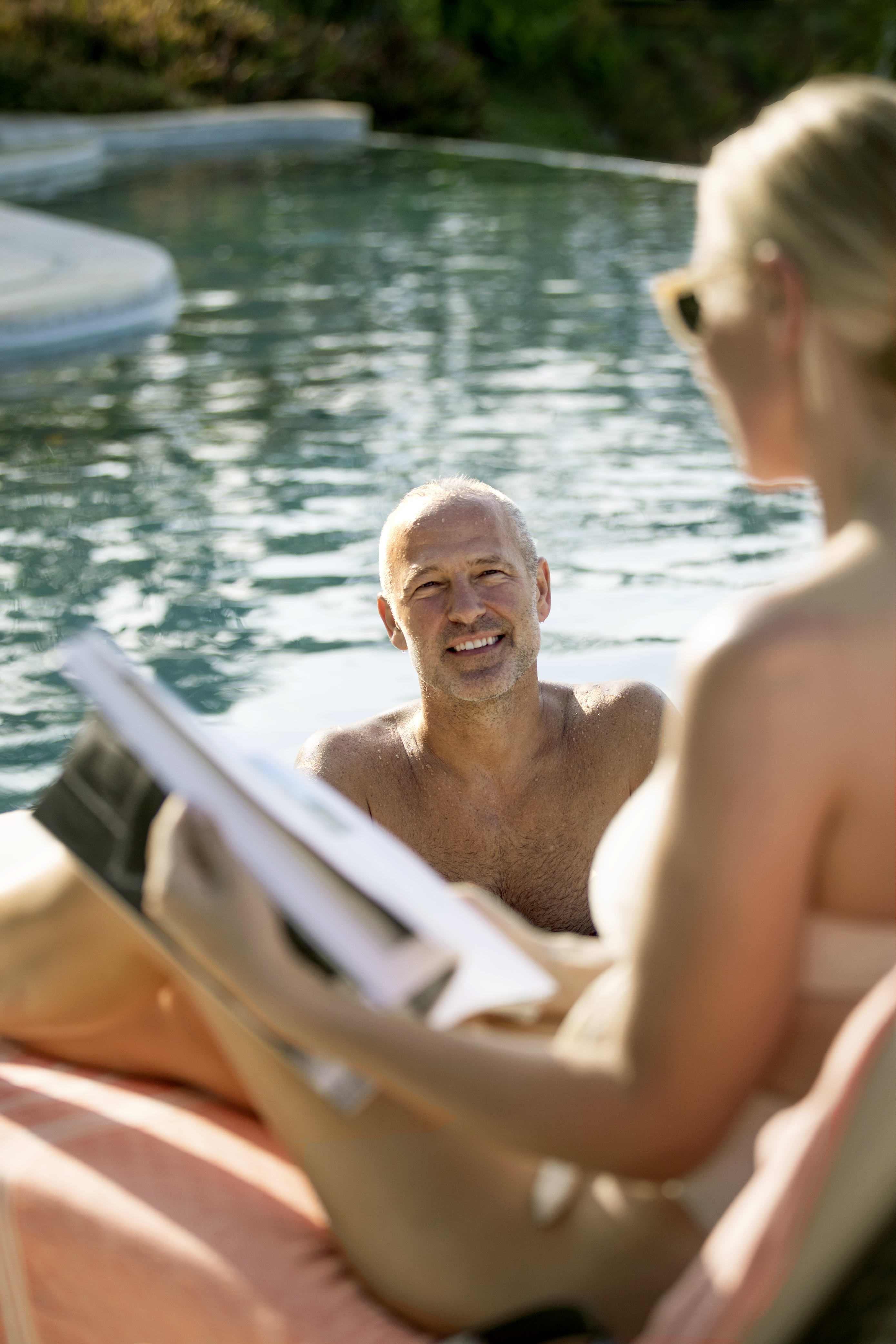 Mother reads a book by the pool while the dad swims.