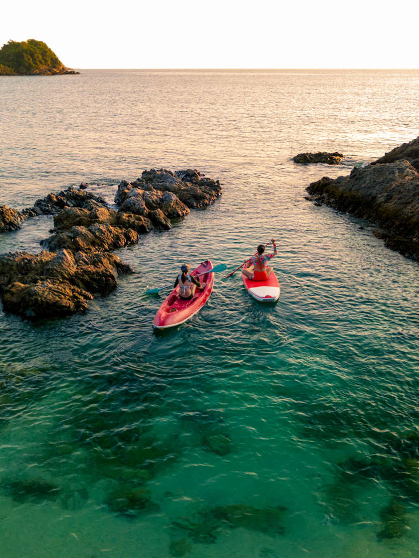 Two people kayaking and paddleboarding through clear turquoise waters, surrounded by rocky outcrops and a distant island.