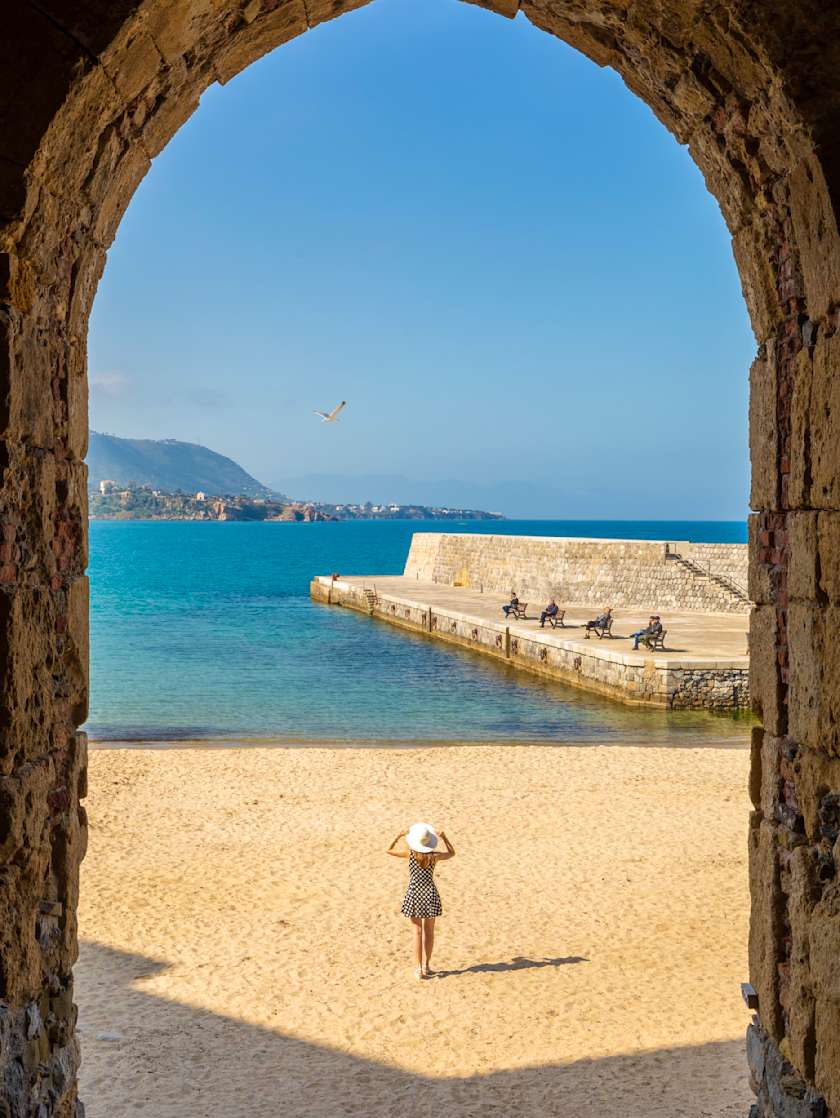 View through a stone archway of a woman standing on a sandy beach, overlooking the blue ocean and distant mountains.