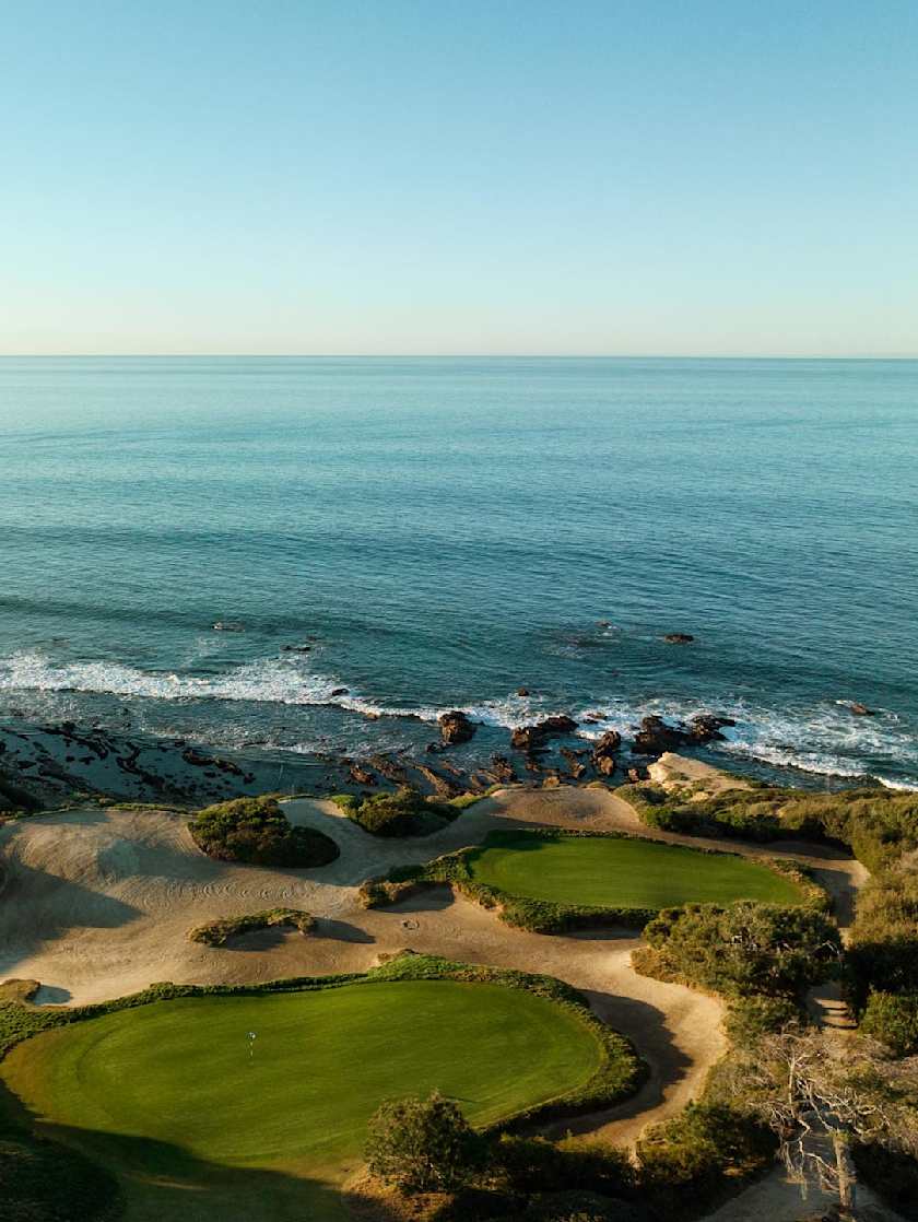 Aerial view of a coastal golf course in Laguna Beach with green fairways, sandy bunkers, and the ocean waves meeting the rocky shoreline.