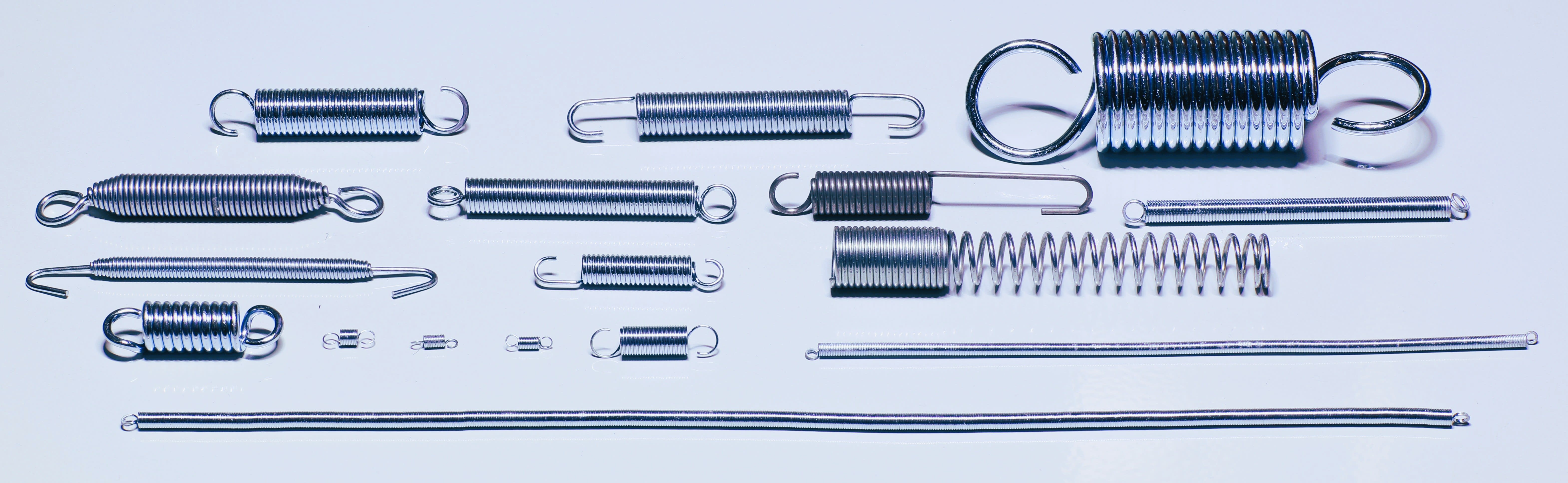 Assortment of new silver metal springs, featuring various sizes of extension springs with hooks and loops, a large compression spring, and two long, thin coiled wires.