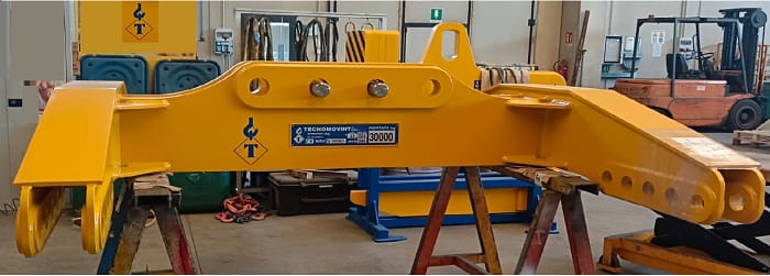 Grande trave bilancino di sollevamento industriale gialla con logo TECNOMOVINT e indicazione di capacità 30000, poggiata su cavalletti in un'officina.