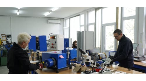 Two individuals performing technical work in a workshop, one operating blue machinery with a control panel, and another examining components with a microscope.