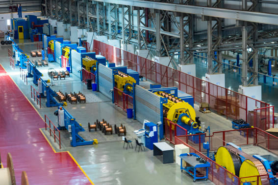 Industrial factory production line with large blue and yellow wire and cable manufacturing machines, spools of copper wire, and red safety barriers.