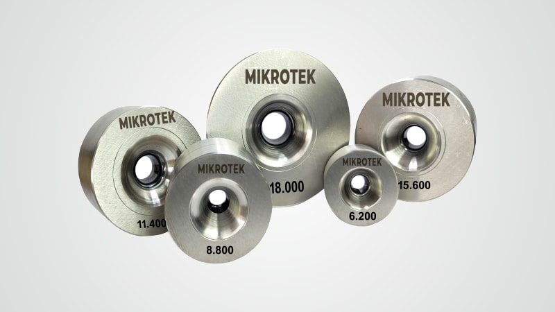 A group of MIKROTEK wire drawing dies of various sizes, made of metal, featuring central conical openings and inscribed numbers like 18.000, 11.400, and 6.200 on their surfaces.