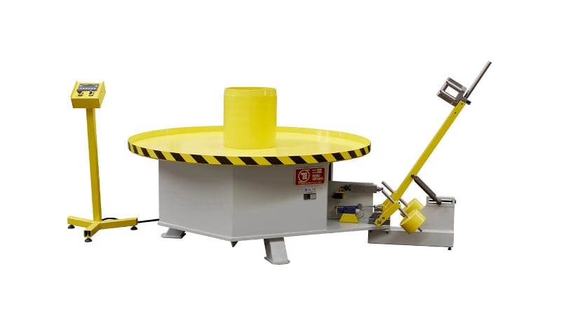 Industrial rotation and impact testing machine for packaging or drums, featuring a yellow turntable with a central cylinder, a control panel, and a roller mechanism on the side.