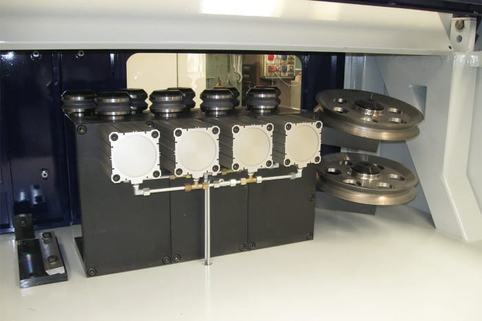 Industrial machine detail featuring five pneumatic actuators on top of control modules, an integrated piping system, and two stacked metal guide pulleys on the right.