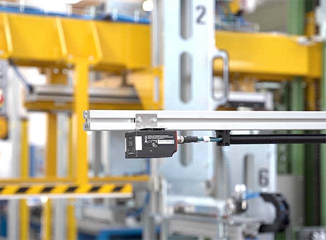 Industrial machine vision camera mounted on a silver rail, overlooking a factory production line with yellow conveyor systems and safety stripes in the background.