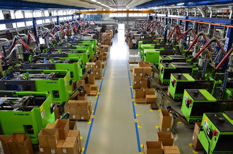 Fabbrica automatizzata con lunghe file di presse ad iniezione verdi ENGEL e-motion 130, equipaggiate con bracci robotici che depositano prodotti in scatole su nastri trasportatori, lungo un ampio corridoio centrale.