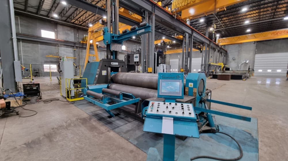 Large blue three-roll plate bending machine with a digital control panel in a spacious industrial workshop, equipped for metal fabrication.