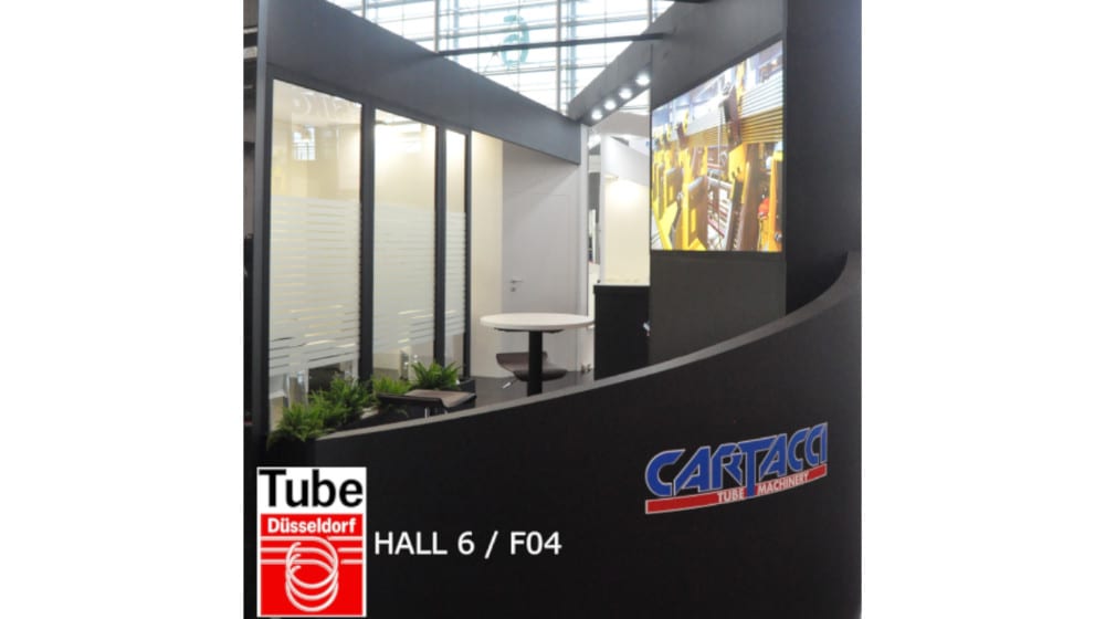 Stand espositivo moderno di Cartacci Tube Machinery alla fiera Tube Düsseldorf (HALL 6 / F04), con area meeting vetrata, bancone curvo nero e schermo che mostra macchinari industriali.