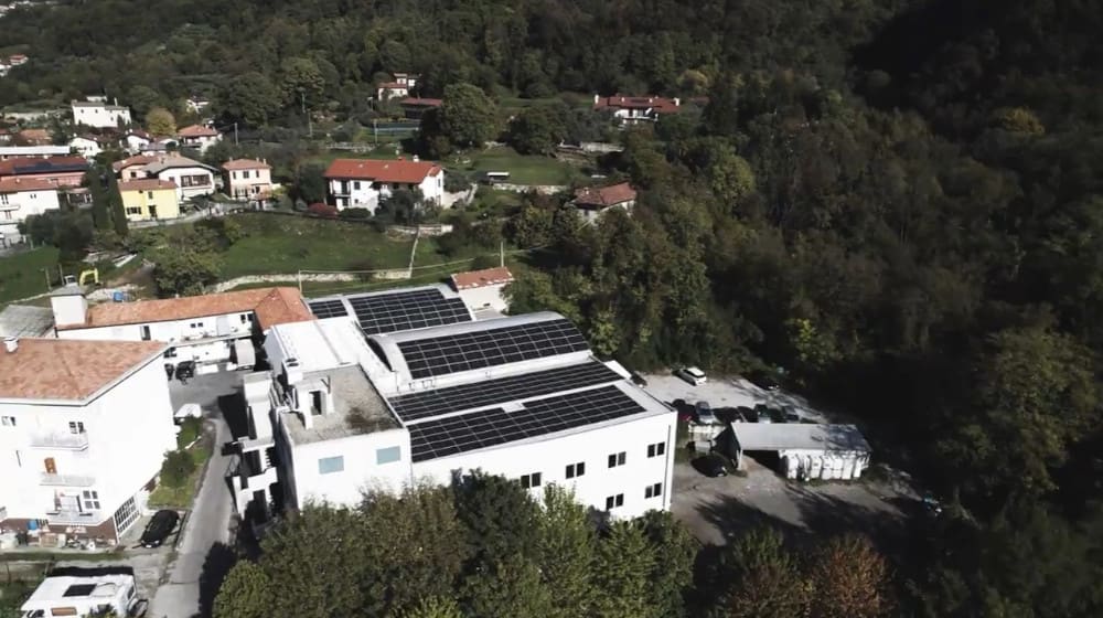Aerial drone shot of a modern commercial building with large rooftop solar panel arrays, nestled among traditional red-roofed homes and dense green forests on a hillside.