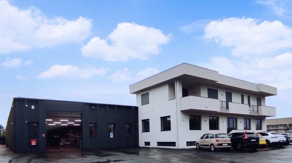 Complesso aziendale moderno con un edificio per uffici bianco a più piani e un capannone industriale grigio scuro con portone aperto, e auto parcheggiate nel piazzale.