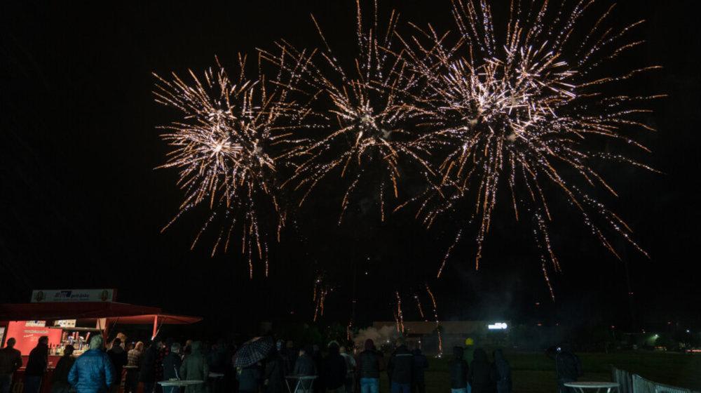 Spektakuläres Feuerwerk am Nachthimmel über einer versammelten Menschenmenge und einem beleuchteten Verkaufsstand bei einer Open-Air-Veranstaltung.