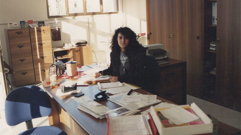 Donna con lunghi capelli ricci scuri seduta alla scrivania di un ufficio, circondata da documenti e attrezzature. Sullo sfondo, mobili in legno chiaro e scuro, con luce solare che illumina la stanza.