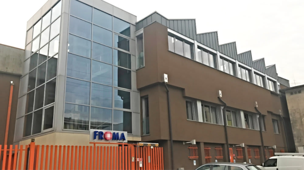 Sede aziendale moderna di FROMA con imponente facciata a vetri, pareti marroni e tetto a shed, visibile dietro una recinzione arancione.