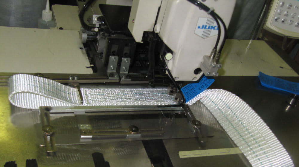 Industrial Juki sewing machine stitching heavy-duty white webbing with green reinforcement lines and blue product labels.