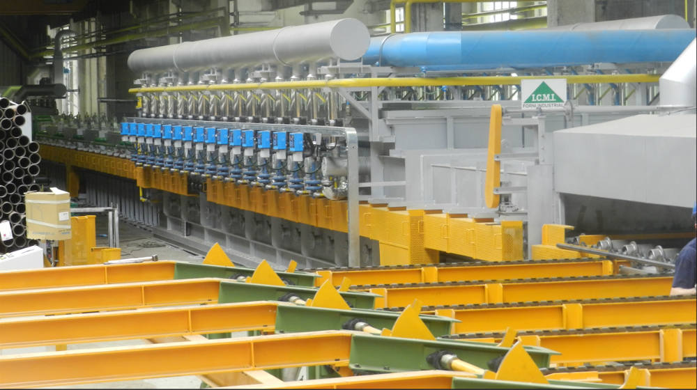 L.C.M.I. industrial machinery with multi-stage conveyor system for pipe processing, featuring blue control units, grey ventilation ducts, and a furnace opening in a manufacturing plant.