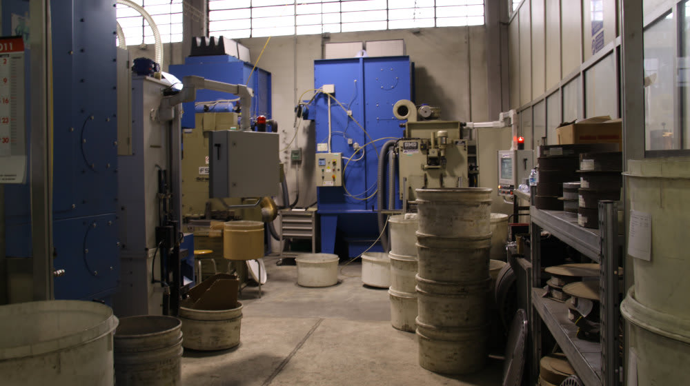 Interno di un'officina industriale con grandi macchinari blu, apparecchiature di lavorazione, contenitori impilati e scaffalature in un ambiente di fabbrica.