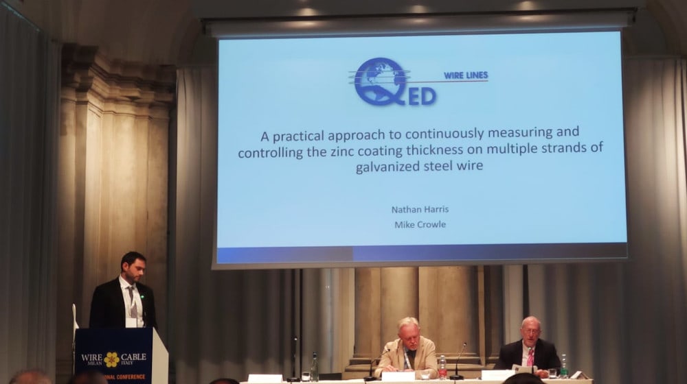 A speaker at the Wire and Cable Italy conference presents on the continuous measurement and control of zinc coating thickness for galvanized steel wire, with QED Wire Lines branding visible on the presentation screen. Two panelists are seated at a table below the screen.