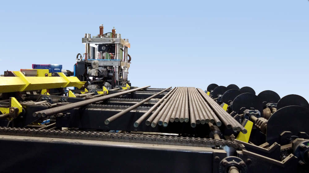 Industrial metal processing line with steel rods on a chain conveyor system and specialized machinery under a clear blue sky.