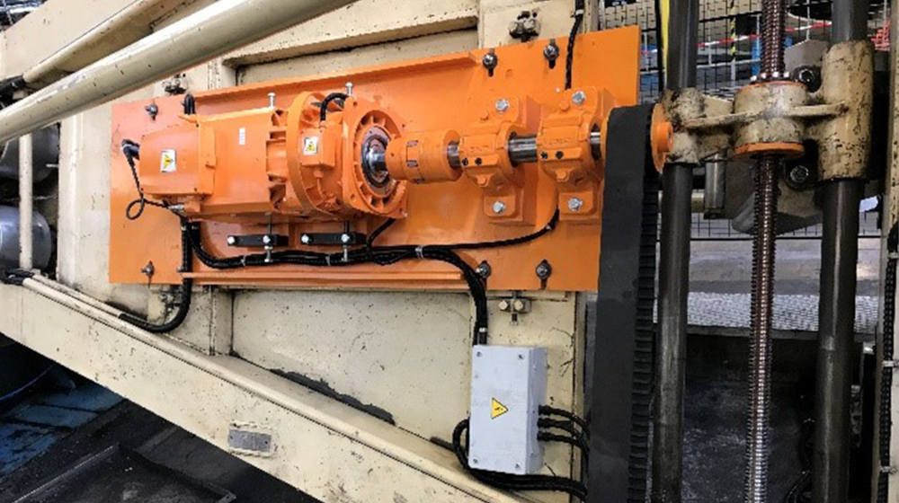 Orange industrial motor and gearbox connected to a timing belt drive system and vertical lead screw on factory machinery.