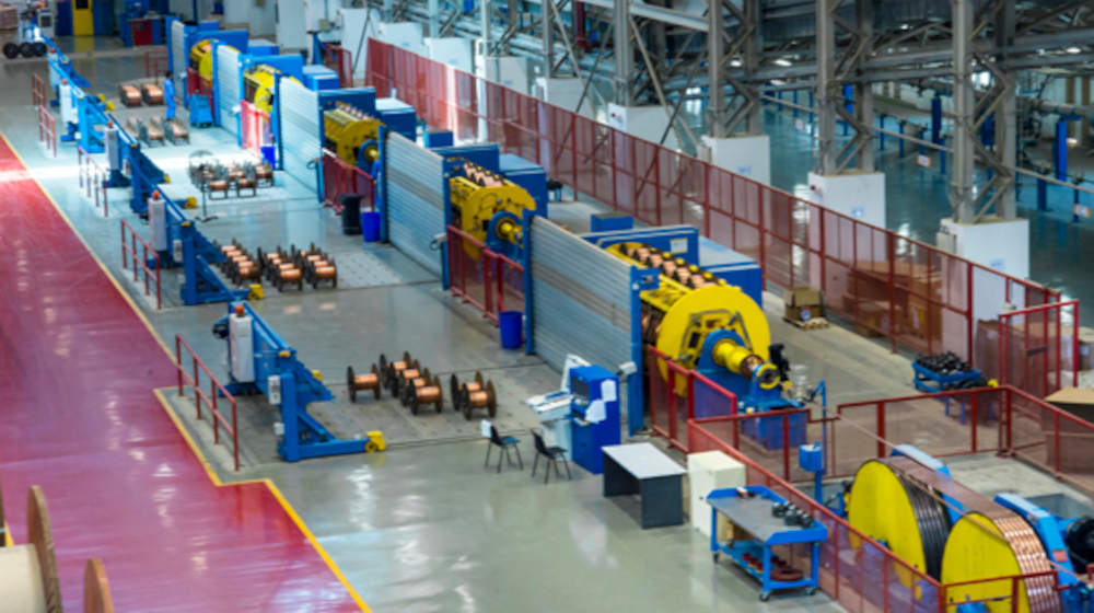 Industrial cable manufacturing facility with an automated production line, featuring large blue and yellow machinery, numerous copper wire spools, and a clear factory floor.