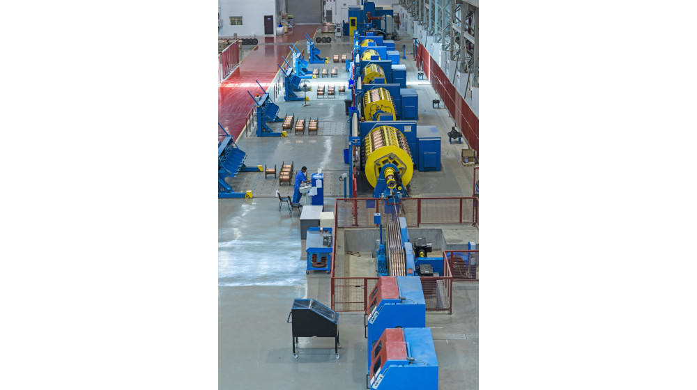 Overhead view of a modern cable manufacturing factory floor, showing a long production line with large blue and yellow industrial machinery, a worker operating a control panel, and numerous spools of material awaiting processing.