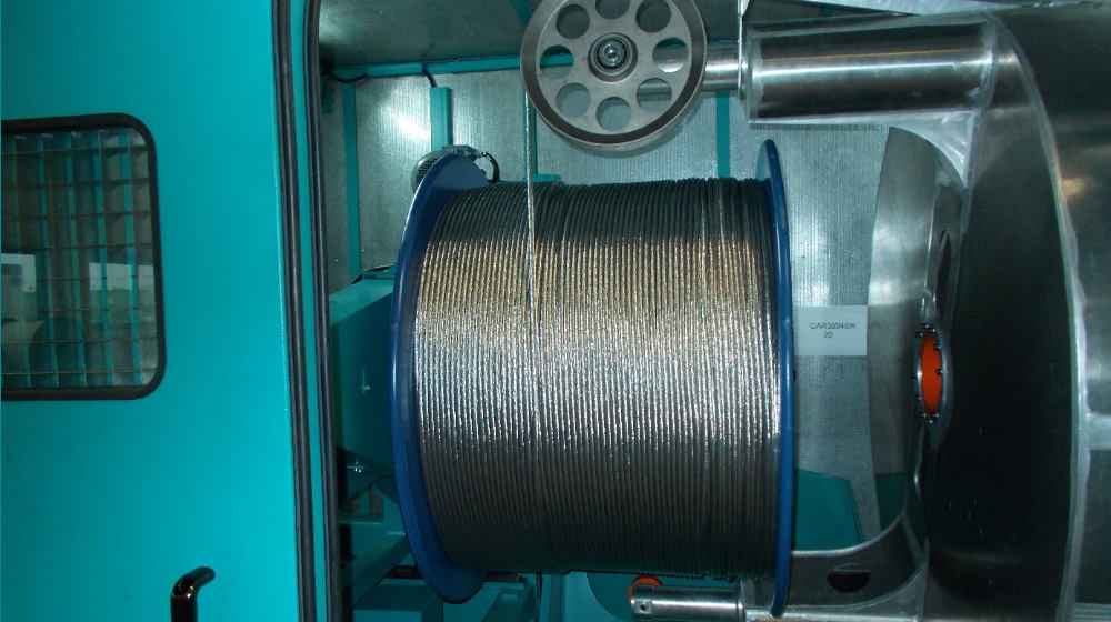 Large industrial spool tightly wound with numerous strands of metallic wire or cable, positioned within a wire manufacturing machine with blue panels and a large pulley.