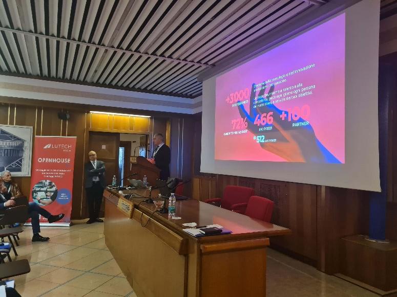 Relatore al podio durante l'evento LUTECH OPENHOUSE, con una presentazione su schermo che mostra statistiche aziendali e una grafica di una mano.