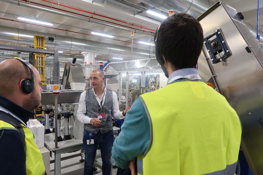Visita guidata in fabbrica: un tecnico con occhiali spiega il funzionamento dei macchinari industriali a due uomini che indossano giubbotti di sicurezza ad alta visibilità e cuffie.
