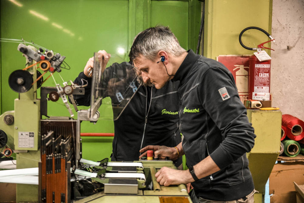 Man in a 'Giorinastri' jacket operating industrial textile machinery, pressing a button while another worker is partially visible behind protective glass. Spools of ribbon and a fire extinguisher are in the industrial background.