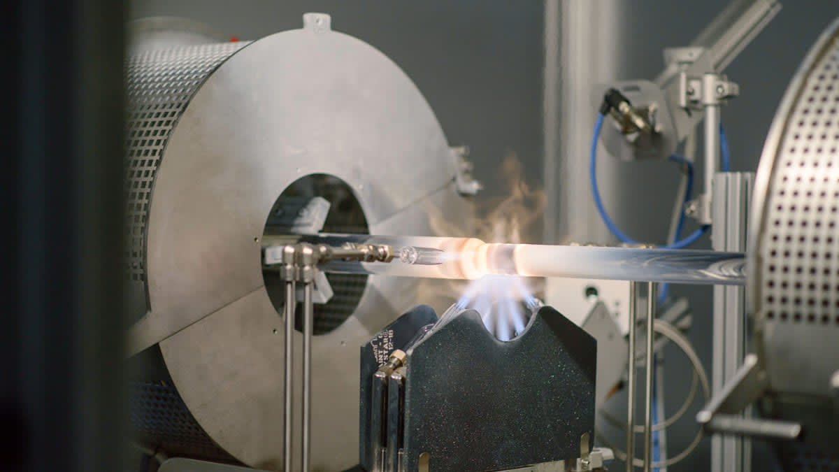 A clear glass tube is intensely heated by a blue flame in a sophisticated industrial or laboratory setting, featuring large rotating metal machinery.
