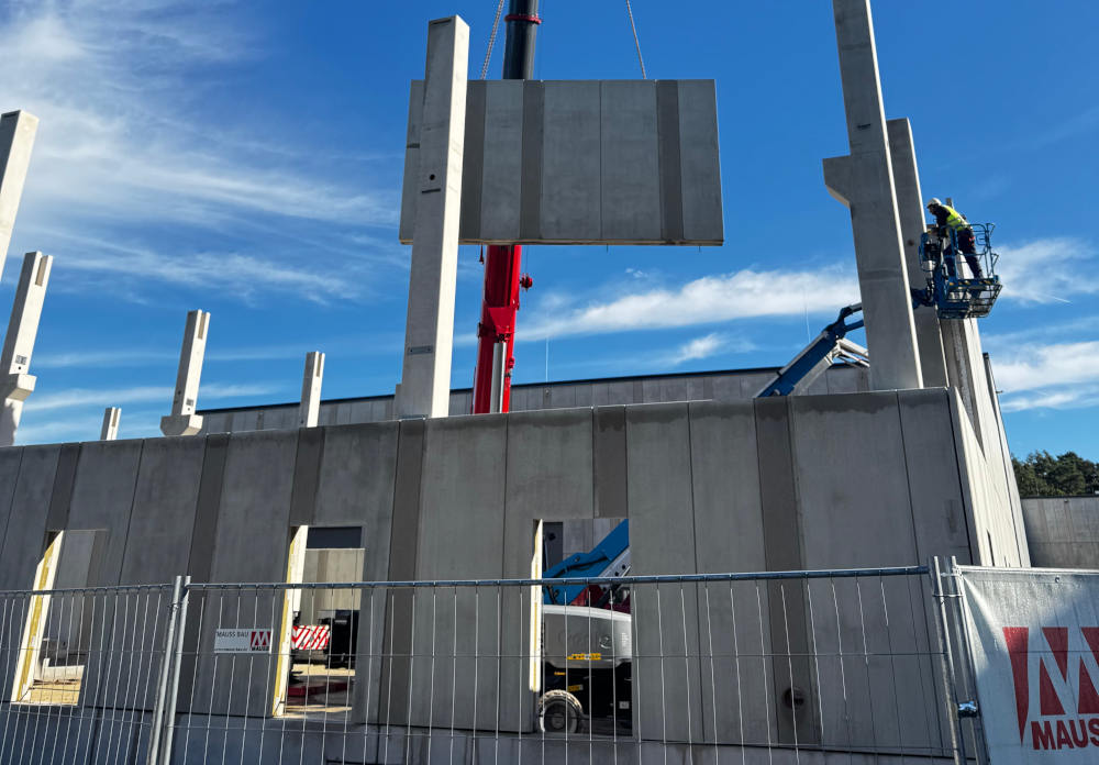 Crane lifting a large precast concrete wall panel onto a construction site, with concrete columns already in place. A worker on an aerial lift stands next to a column, and a Mauss Bau banner is on a fence in the foreground.