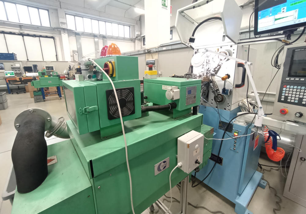 Impianto industriale automatizzato per la lavorazione e formatura di precisione, composto da un'unità di processo verde in primo piano e una macchina CNC blu con monitor di controllo e meccanismo di avvolgimento, all'interno di un'officina moderna.