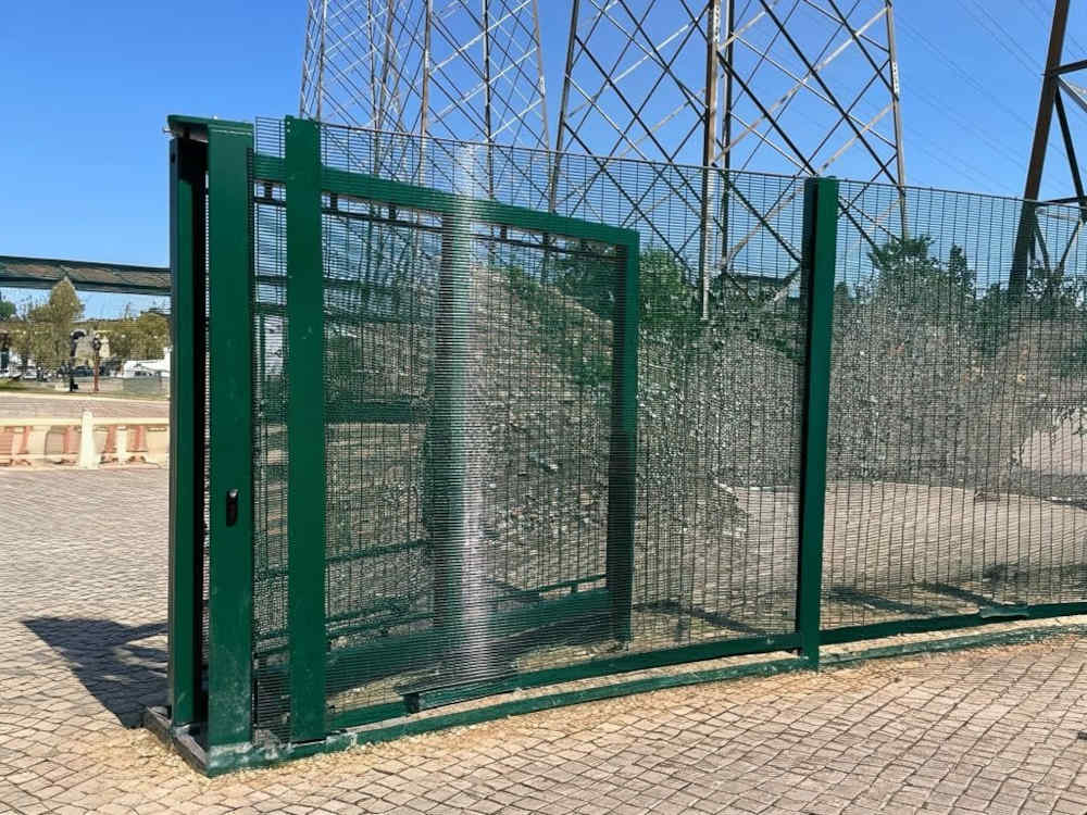 Dark green high-security mesh fence and partially open gate on a paved area, with large electricity pylons in the background under a clear blue sky.